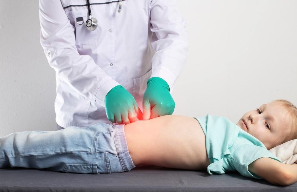 Pediatrician in a white coat with green gloves palpating a child's abdomen during an exam on a medical table.