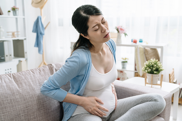 Picture2 - Dr Ksheetij Kothari Pregnant woman sits on a sofa at home, hands on her belly and lower back, showing discomfort or backache.