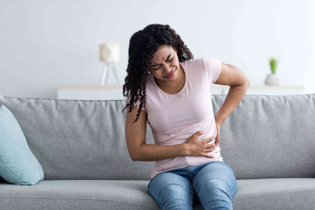 Person in a pink sweater holds their stomach with one hand while gripping a glass of dark red drink in the other, suggesting stomach discomfort or nausea.