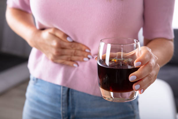Person in a pink sweater holds their stomach with one hand while gripping a glass of dark red drink in the other, suggesting stomach discomfort or nausea.